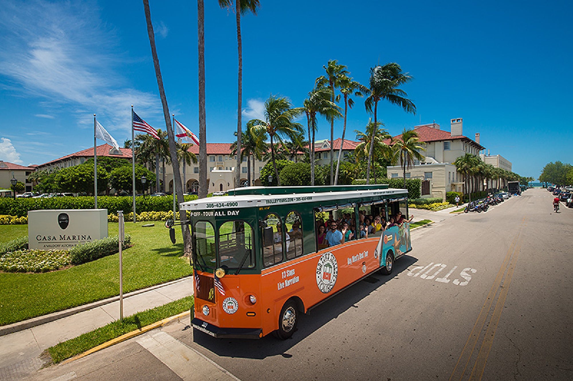 An orange and green tour trolley drives past the Casa Marina hotel, with palm trees, flags, and a clear blue sky in the background.
