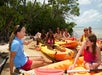 A group of people in kayaks listens to an instructor on a sandy shore near trees and water.