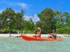 Two people kayaking in a bright orange kayak on clear water, with a backdrop of green trees and a blue sky.