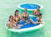 Five women in swimsuits sit in an inflatable pool float on clear water, holding drinks and smiling at the camera. Another empty float is visible in the background.