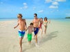 A group of five people enjoy walking through shallow water on a beach under a clear blue sky.
