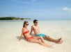 A couple in swimwear relaxes on a sandy beach, sitting in shallow, clear water with a sunny sky and green foliage in the background.
