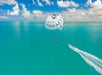 A parasailer is being towed by a boat over clear turquoise water under a blue sky with clouds.
