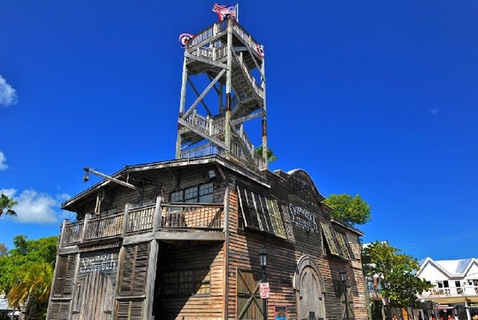 A weathered wooden building with a tall observation tower, American flag on top, and clear blue sky in the background.