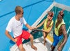 Three people in swimwear and snorkeling gear are on a boat. Two are in the water, holding flippers, while one sits on the boat, wearing a white shirt and red shorts, smiling at them.