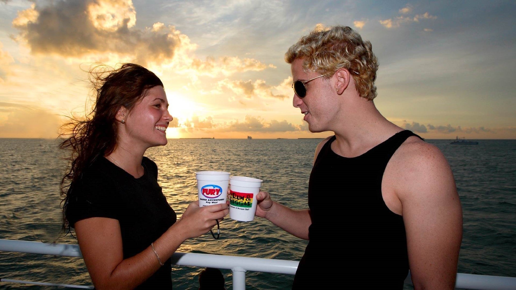 Two people in casual attire enjoy drinks on a boat at sunset, smiling at each other with the ocean in the background.