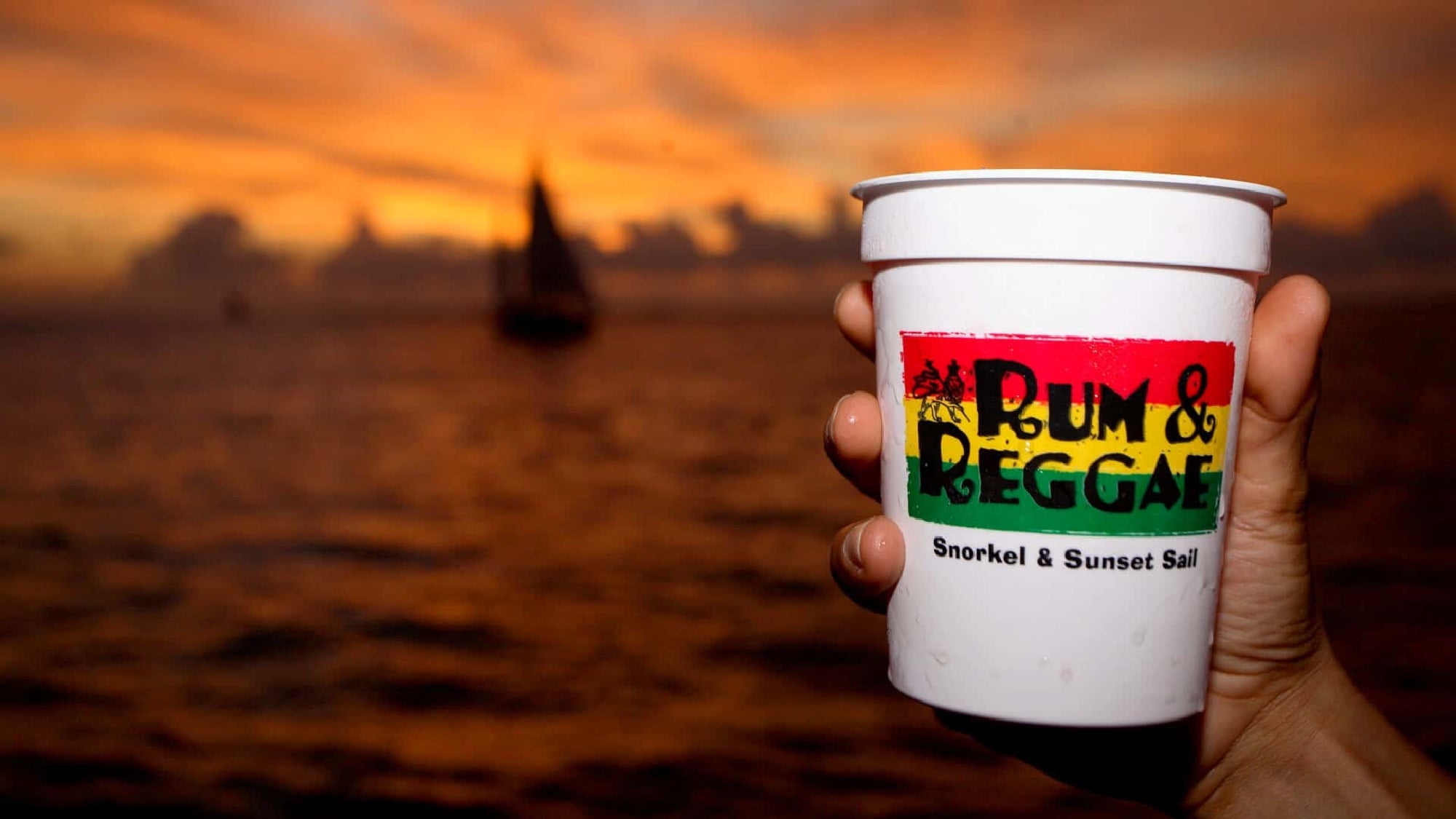A hand holds a white cup with "Rum & Reggae Snorkel & Sunset Sail" printed on it. A sunset and sailboat are visible in the background.