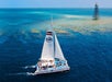 A sailboat navigates clear blue waters near a small lighthouse structure, with coral reefs visible beneath the surface.