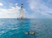 Two people snorkel in the foreground while a large sailboat is visible in the background on a calm blue ocean under a partly cloudy sky.