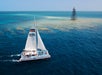 A catamaran sails near a coral reef with a lighthouse in the background on a clear day.