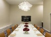 A modern meeting room with a long white table, brown chairs, a central flower arrangement, and a display screen reading "Kimpton Hotels & Restaurants." A white ceiling light fixture hangs above.