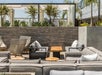Modern outdoor patio with gray cushioned seating, a wooden table, and woven chairs. Surrounded by potted plants and stone walls, under a clear blue sky.