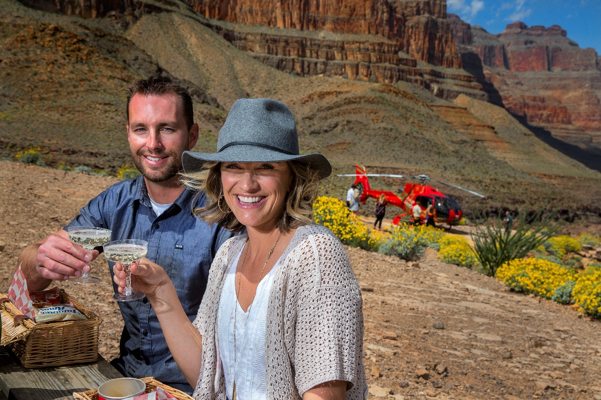 A couple enjoys drinks at a picnic spot in a canyon landscape, with a red helicopter and yellow flowers in the background.