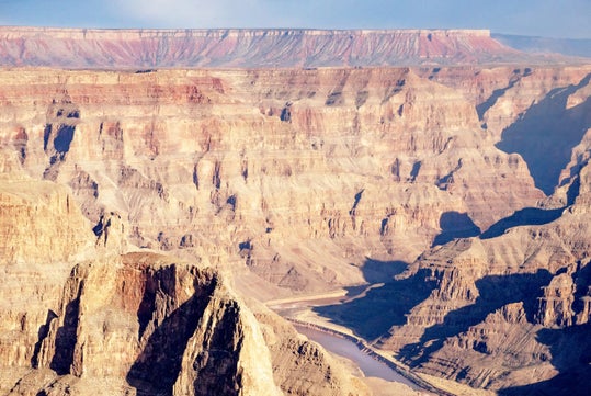 Aerial view of the Grand Canyon showing layers of colorful rock formations and a river winding through the deep, expansive landscape.