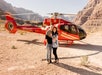 A couple stands embracing in front of a red helicopter on a rocky terrain with mountains in the background.
