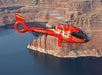 A red helicopter with the number 23 flies over a vast canyon landscape with cliffs and water below.
.