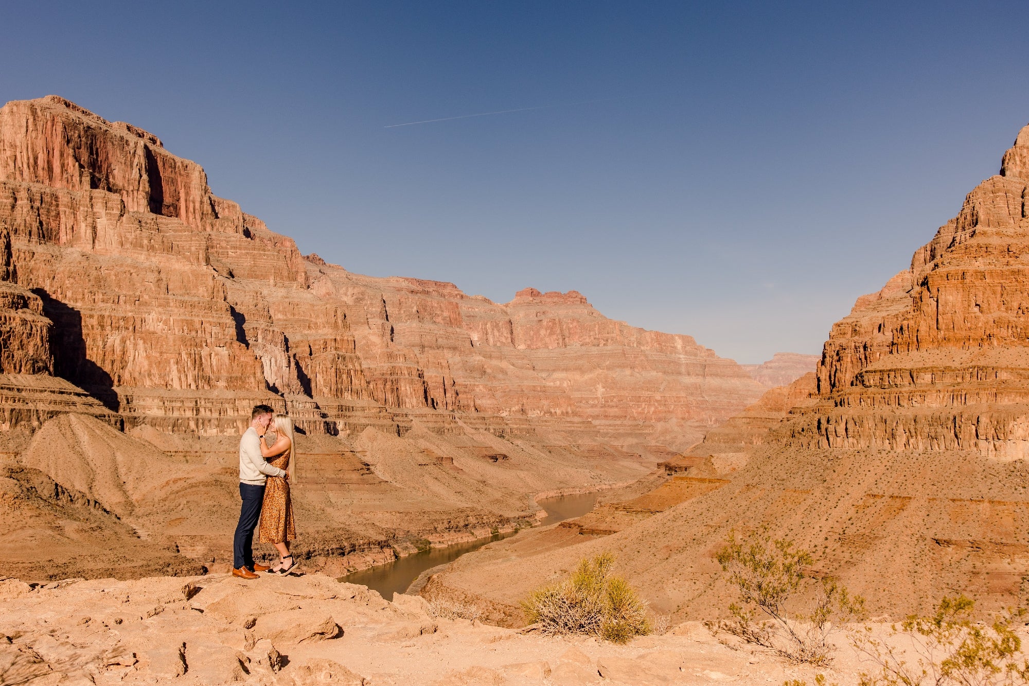A couple embraces on a cliff overlooking a canyon with a river beneath a clear blue sky.