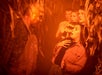 A group of people look startled as a man in overalls with face paint confronts them in a cornfield with orange lighting.