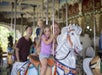 A family of four rides on a carousel; a young girl sits on a white horse in front, smiling, with her parents and a younger boy behind her.