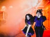 Two people in purple and gold jester costumes stand in a brightly lit, smoky area; one smiles at the camera while the other covers their face with their hand.