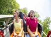 Two teenagers in the front row of a roller coaster ride, gripping the safety bar and expressing excitement and fear, with other riders visible behind them. Trees and sky are in the background.