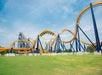 A large, orange and blue steel roller coaster with multiple loops and curves stands on a grassy field under a partly cloudy sky.