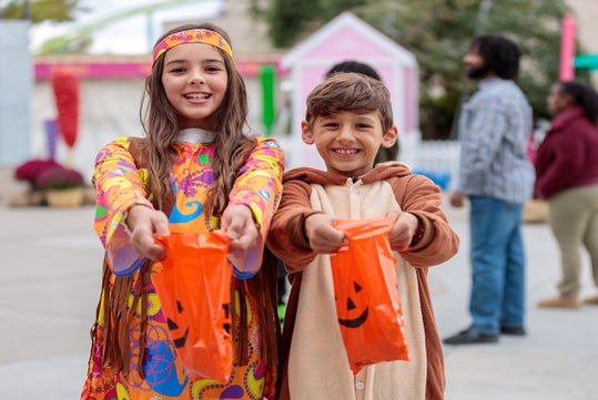 Two children in costumes holding orange Halloween treat bags, smiling at the camera outdoors, with people and buildings in the background.