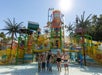 Six children in swimsuits stand and pose in the shallow water at a colorful outdoor water park with slides and palm tree structures under a bright sun.