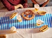 Two people sit at a table with a large pretzel, three hot dogs, and four cups of beer on a blue and white checkered tablecloth.