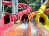 A person emerges from a bright orange water slide, arms raised, with splashes of water around, at an outdoor water park with multiple colorful slides in the background.