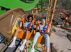 Two people riding a suspended roller coaster, holding safety guards, smiling and laughing with their legs dangling above the ground at an amusement park.