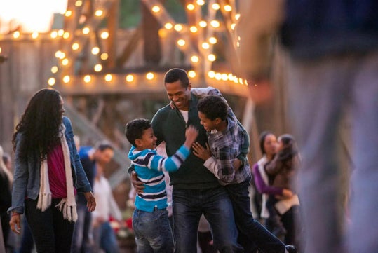 A man smiles while hugging two children outdoors; a woman walks nearby. String lights and a wooden structure are in the background.