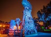 A wooden windmill structure decorated with artificial cobwebs and colorful lighting stands in front of rustic buildings at night.