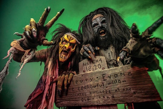 Two people in scary monster costumes pose with outstretched hands, holding a wooden sign with text, against a green-lit background.