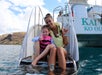 A woman and a young girl in swimsuits and life jackets sit on a boat slide, smiling and making shaka hand signs, with a boat and rocky hills in the background.