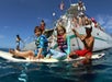 Two children in life jackets sit on a surfboard in the water next to a boat with several people on board under a clear blue sky.