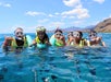 Five people wearing snorkel gear and life jackets float in clear blue water near a mountainous shoreline under a partly cloudy sky.