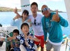 A group of five people, including children and adults, smiling and making shaka hand signs on a boat with blue water and coastline in the background.