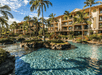 A resort pool with clear blue water is surrounded by palm trees and a large building under a bright blue sky.