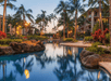 A serene tropical resort pool at sunset, surrounded by palm trees and buildings. Fiery torches light the path, reflecting on the calm water.