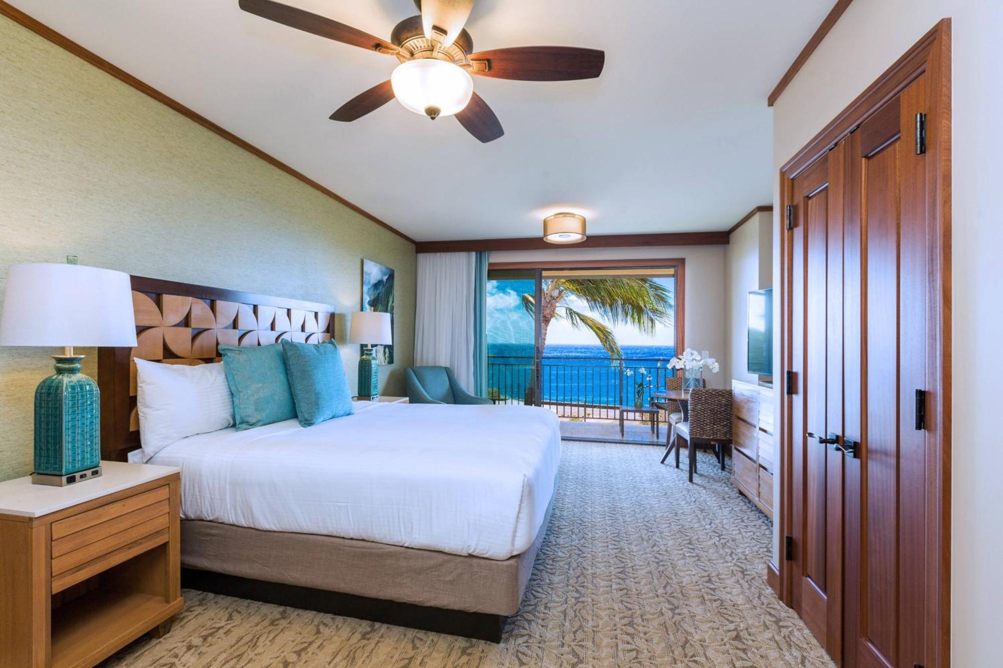 Hotel room with a king-sized bed, nightstands, ceiling fan, desk, dresser, and a balcony overlooking the ocean and palm trees.