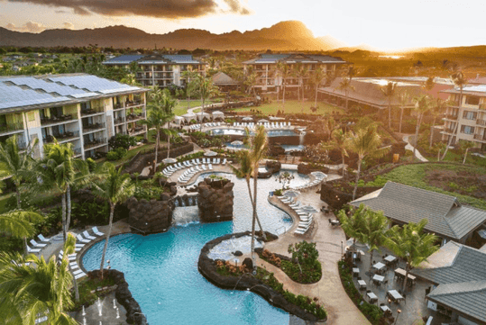 Aerial view of a resort with multiple pools, sun loungers, and palm trees surrounded by buildings, against a backdrop of mountains and a sunset sky.