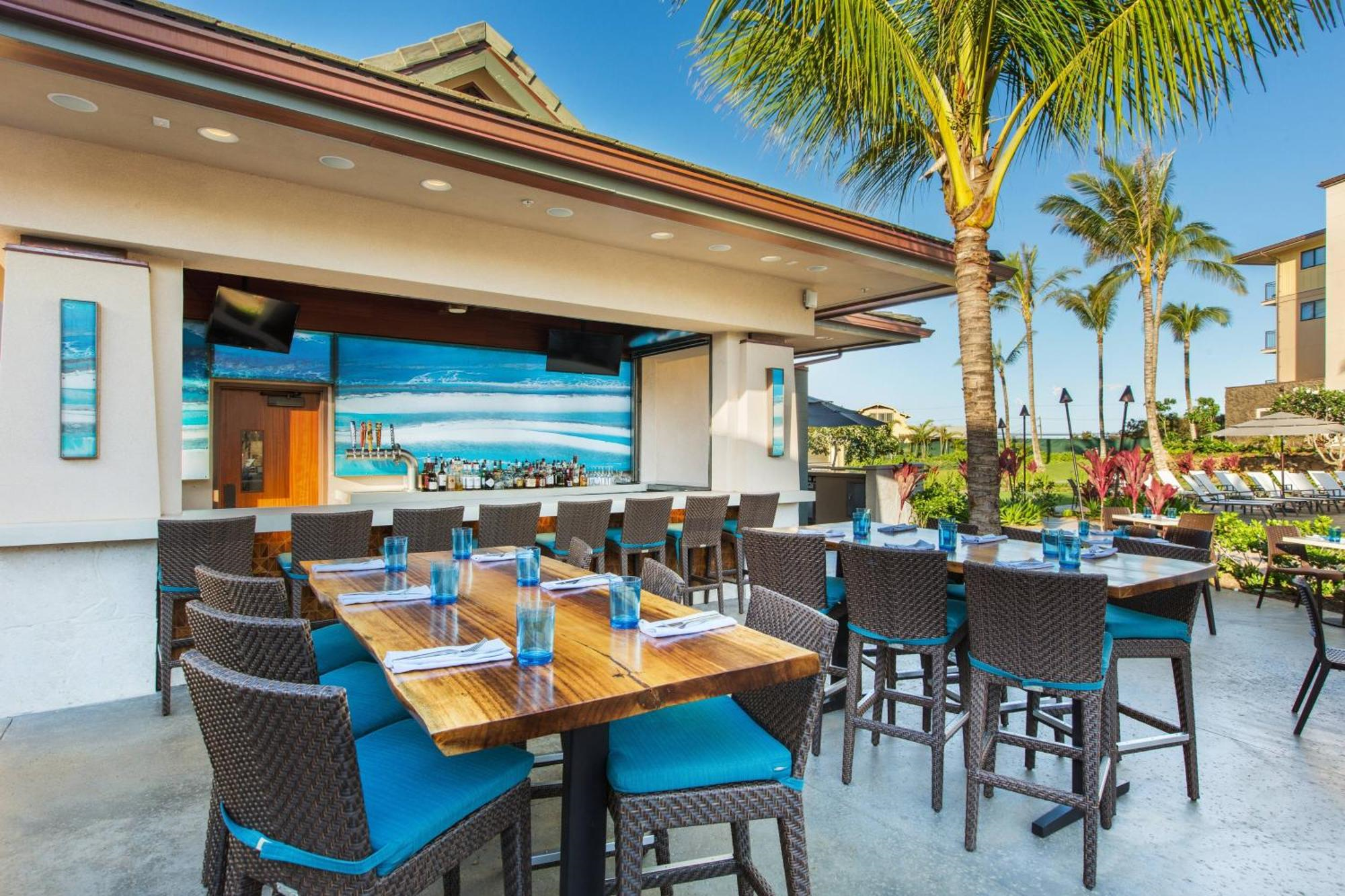 Outdoor restaurant patio with wooden tables and wicker chairs, place settings with blue glasses, palm trees, and a mural of an ocean scene on the wall.