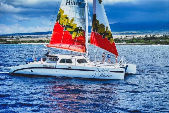 A white and red catamaran named "Spirit of Aloha" with people on board sails on blue water near a coastline under a partly cloudy sky.