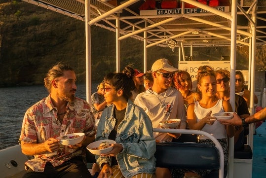 A group of people sitting on a boat enjoying food, with a rocky shoreline visible in the background during sunset. Some are holding bowls while engaging in conversation.
