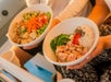 Two people holding bowls of food; one with diced tofu, vegetables, edamame, and carrots, the other with rice, diced fish, seaweed salad, and roe.