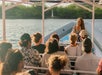 A group of people sit on a boat with a woman standing at the front, talking. The boat is on water with greenery in the background.