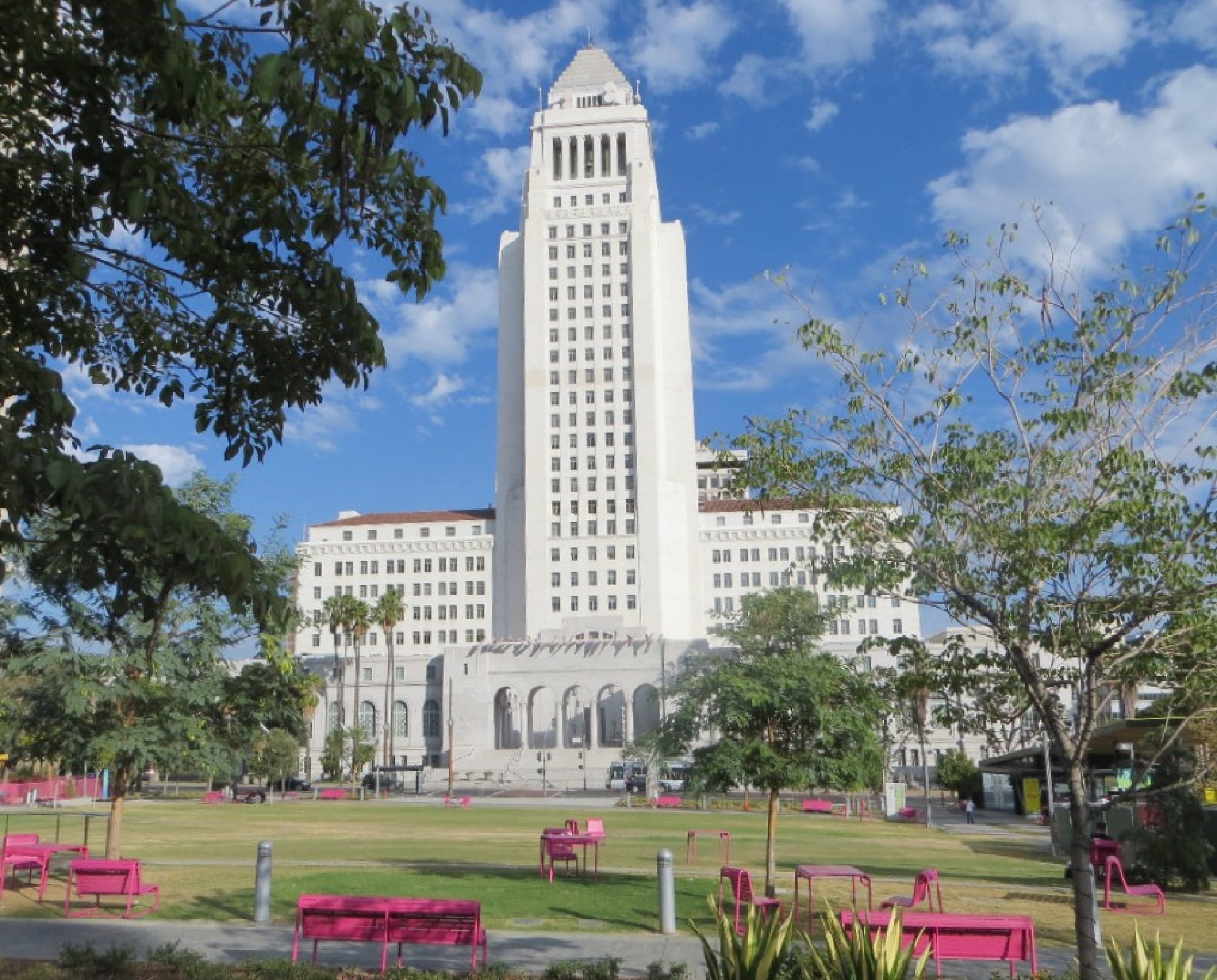 A tall, white tower building stands against a blue sky with some clouds. In the foreground, there is a green lawn with pink benches and trees.