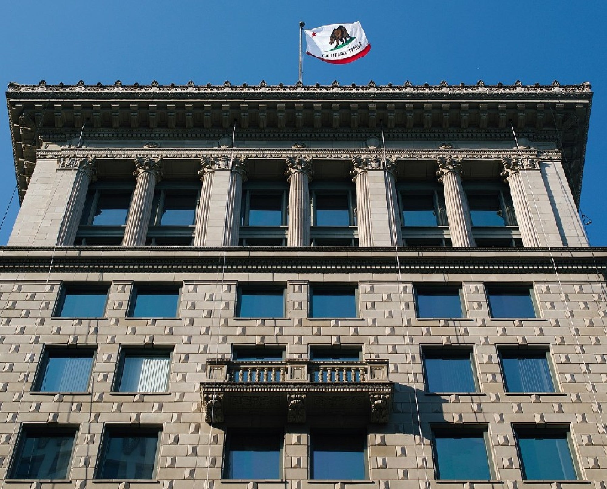 A multi-story building with multiple windows and a balcony centered on the façade. The California state flag is flying atop the building against a clear blue sky.