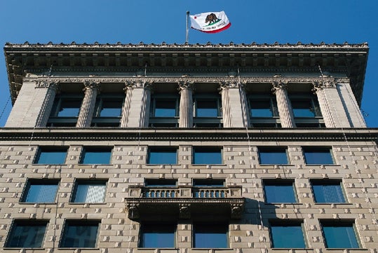 A multi-story building with multiple windows and a balcony centered on the façade. The California state flag is flying atop the building against a clear blue sky.